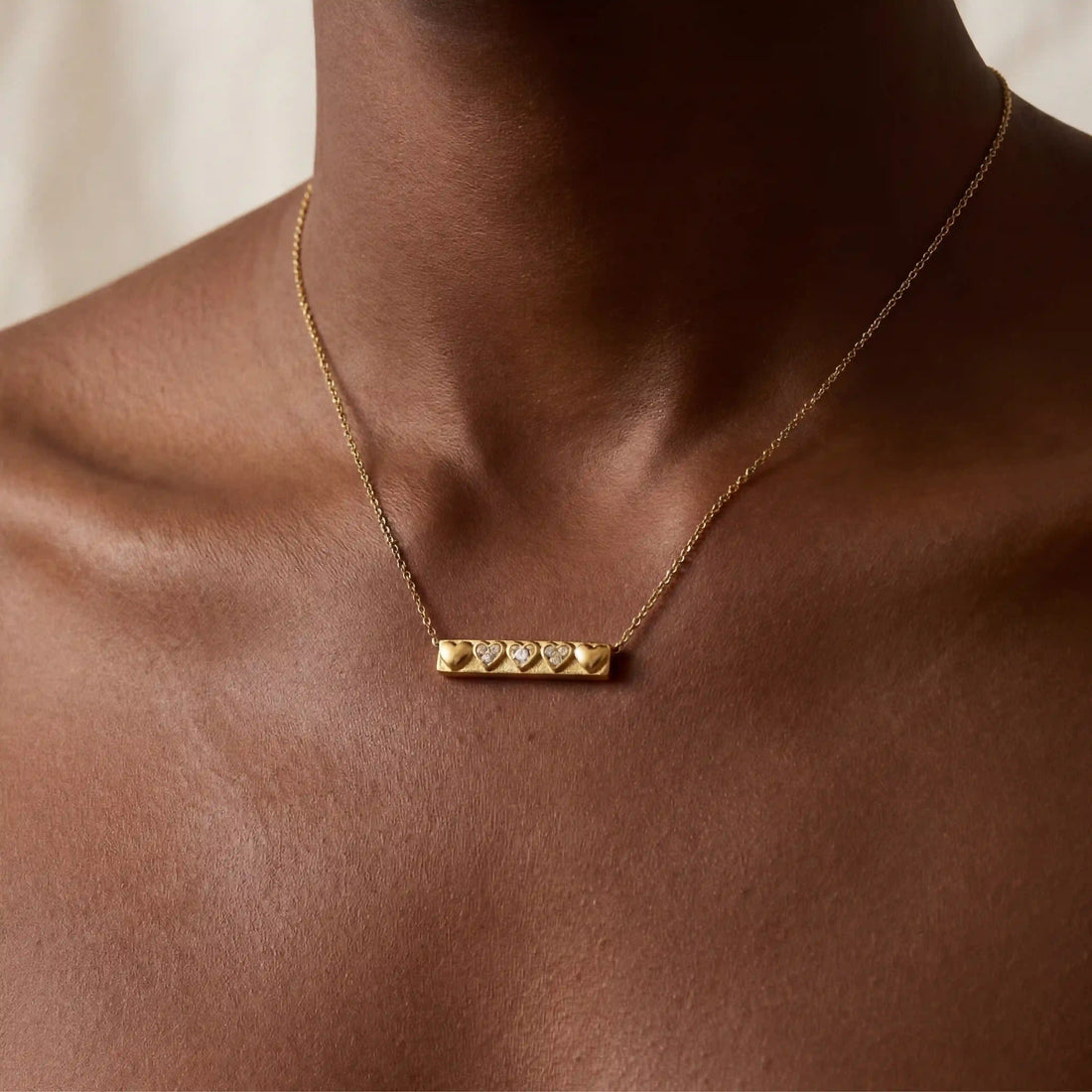 Close up of woman with dark skin wearing gold bar necklace with row of heart details.