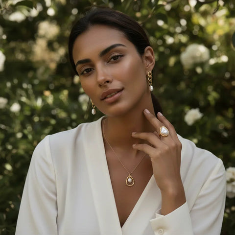 A beautiful model in a lush garden setting, showcasing a complete Luminova Jewelry look. She is wearing The Theia baroque pearl statement ring, a matching sculptural pearl pendant necklace, and elegant gold link and pearl drop earrings.