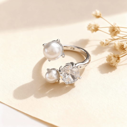 A silver version of The Celestial Ring displayed side-by-side on a neutral, textured brown surface. Each adjustable open ring features one large CZ crystal and two pearls in an asymmetrical design.