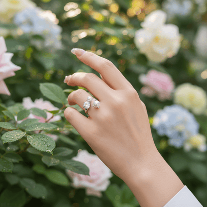 A model's hand, wearing The Celestial Ring, delicately touching the soft petals of a pink peony. The image contrasts the soft, organic beauty of the flower with the sparkle of the crystal and the luster of the pearls.